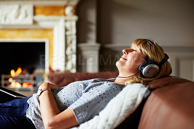 Buy stock photo Shot of a mature woman lying on her sofa listening to music on headphones with a fire in the background