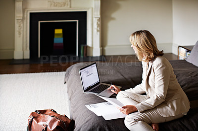 Buy stock photo Shot of a mature businesswoman sitting on a hotel bed using a laptop and reading paperwork
