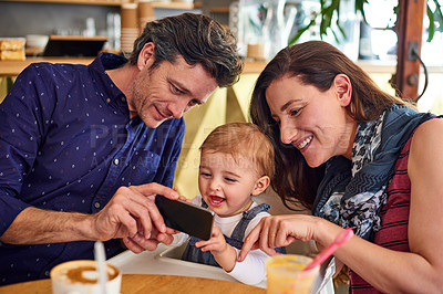 Buy stock photo Shot of a mother and father showing their baby girl photos on a phone while sitting in a cafe