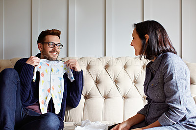 Buy stock photo Shot of a young man showing his pregnant wife baby clothes on their sofa