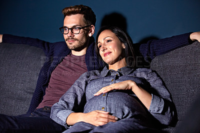 Buy stock photo Shot of a pregnant woman and her husband sitting on the sofa watching television in the evening