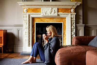 Buy stock photo Shot of a mature woman sitting against her sofa drinking tea while wrapped in a blanket