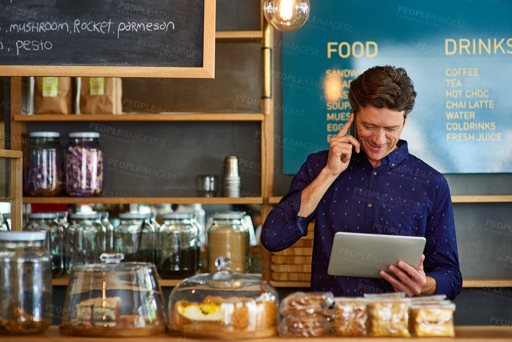 Buy stock photo Shot of a man talking on the phone and using a digital tablet while standing behind a counter in a cafe