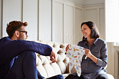 Buy stock photo Shot of a young pregnant woman showing her husband baby clothes on their sofa