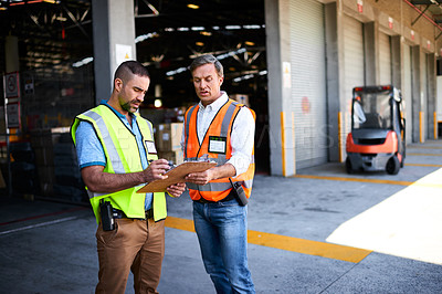 Buy stock photo Shot of two workers talking together over a clipboard while standing at the entrance of a warehouse