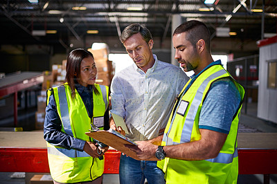 Buy stock photo Shot of three workers talking together over a clipboard inside of a warehouse