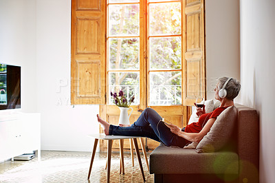 Buy stock photo Shot of a mature woman sitting on her sofa listening to music on a digital tablet and drinking wine