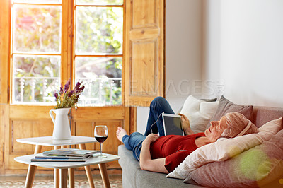 Buy stock photo Shot of a mature woman lying on her sofa listening to music on a digital tablet and drinking wine