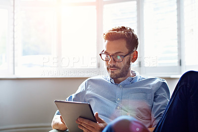 Buy stock photo Shot of a young man sitting on his sofa at home using a digital tablet