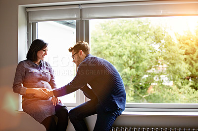 Buy stock photo Shot of a man touching his pregnant wife's stomach while sitting on a window sill at home