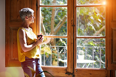 Buy stock photo Shot of a mature woman standing by a window at home drinking a cup of coffee