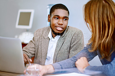 Buy stock photo Shot of two young colleagues talking together while working on a laptop at a desk in an office