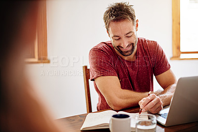 Buy stock photo Shot of a man writing in a notebook and using a laptop while sitting at a table in an office
