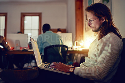 Buy stock photo Shot of a young designer working on a laptop in an office late in the evening