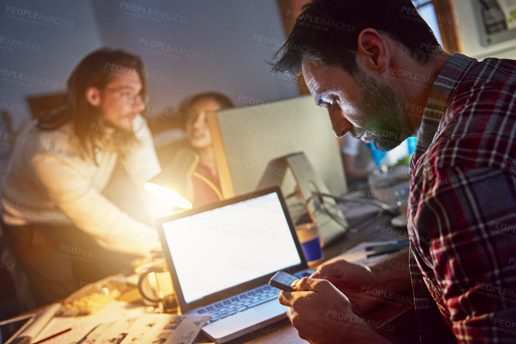 Buy stock photo Shot of a man using a cellphone and working on a laptop in an office in the evening