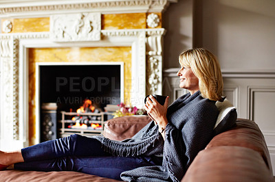 Buy stock photo Shot of a mature woman sitting on her sofa drinking tea while wrapped in a blanket