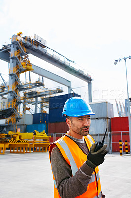 Buy stock photo Shot of a young man in workwear standing outside on a large commercial dock talking on a walkie-talkie