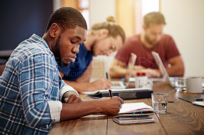 Buy stock photo Shot of a young man writing in notebook while sitting at a table in an office with colleagues in the background