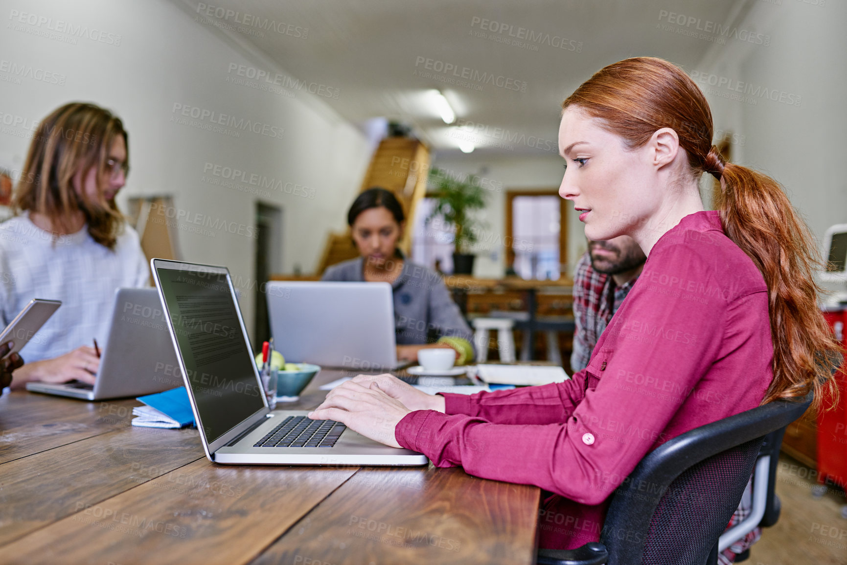 Buy stock photo Shot of a young woman using a laptop at a table in an office with colleagues in the background