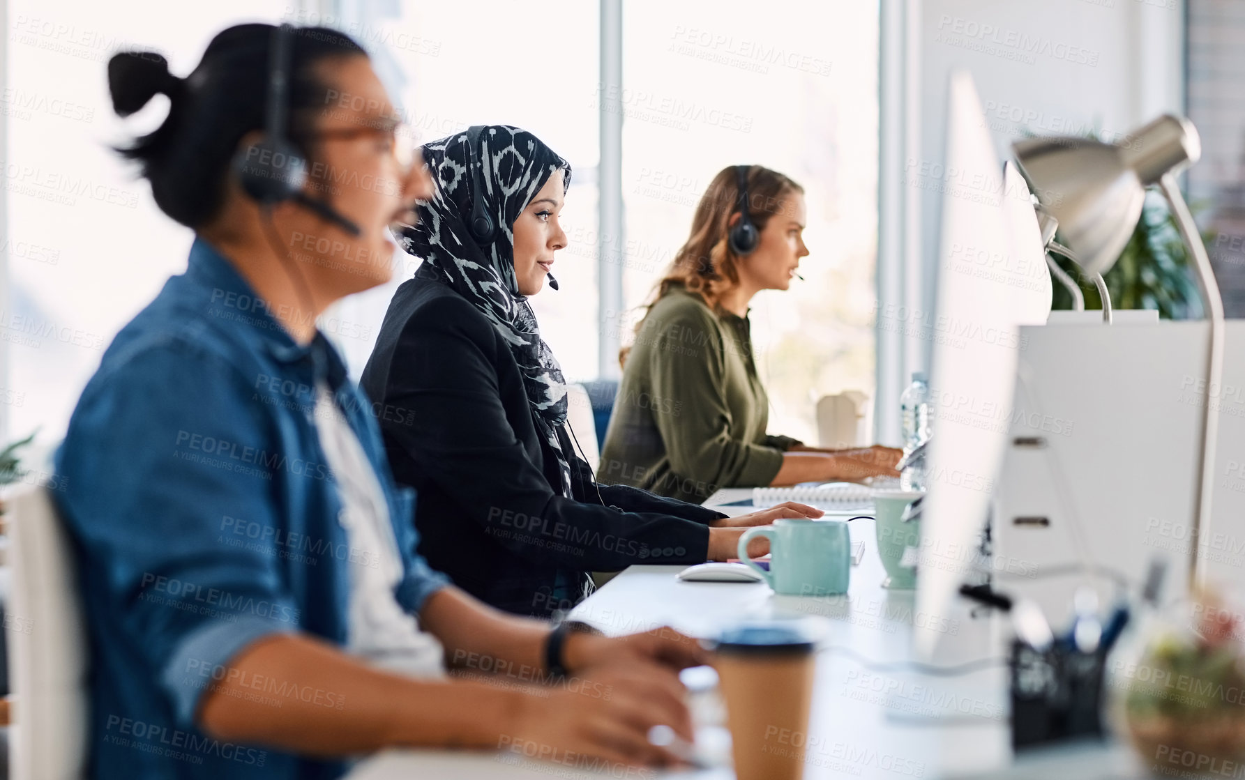 Buy stock photo Shot of a group of young call centre agents wearing headsets and working on their computers in an office