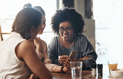 Buy stock photo Shot of attractive young women sitting together and smiling while looking at a cellphone in a coffee shop