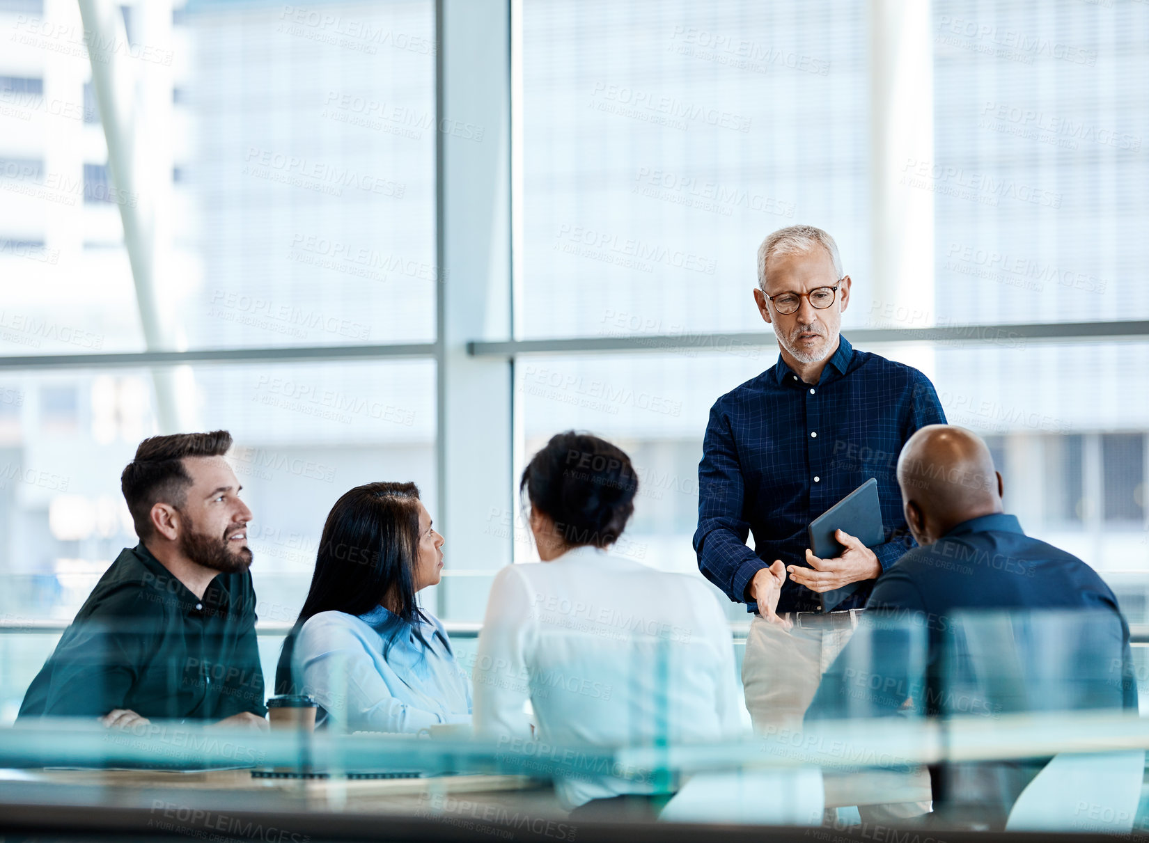 Buy stock photo Shot of a mature businessman using a digital tablet during a meeting with colleagues at work