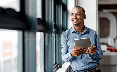Buy stock photo Shot of a young businessman using a digital tablet while standing at a window in an office