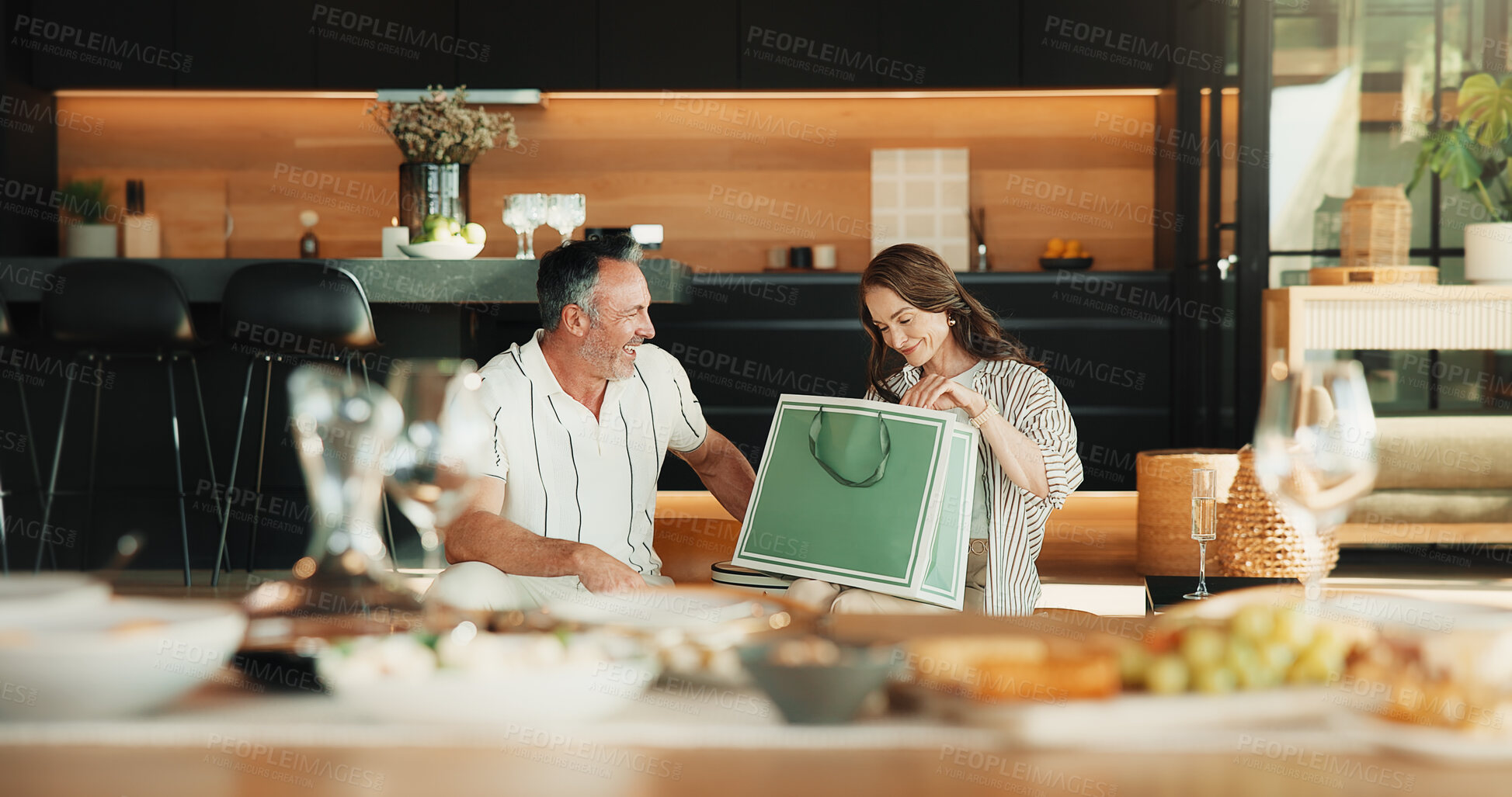 Buy stock photo Love, gift bag and mature couple in home, laugh and gratitude for anniversary celebration with smile. Man, woman and appreciation with present, care or marriage milestone with funny partner at lunch