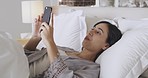 One beautiful mixed race woman sending a text using her phone while lying in bed. Young hispanic woman reading something interesting on social media or replying to a message