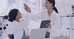 Two scientists high five while using a digital tablet in a laboratory. Happy female doctors celebrate an achievement after making a scientific discovery. Successful research results in advanced lab