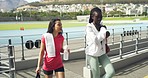 Two young diverse female athletes getting ready to run together at a stadium. Happy sporty friends and teammates arriving at a field to train. Fit woman preparing for a workout at a sports club