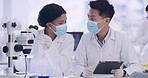 Two medical scientists using a tablet and microscope to check virus samples inside a research facility. Specialists enter data into a system and analyze it for breakthrough, discovery and innovation