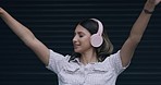 Woman listening to music on headphones and dancing with her arms raised while isolated against a black background. One carefree, fun, expressive woman with increased serotonin enjoying loud audio 