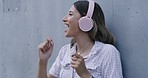 Trendy young woman wearing headphones and listening to music while dancing and singing outside. Cheerful young female with a playful attitude having fun while against a grey wall with copyspace