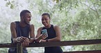 Two joggers using their phone in a park and looking at a fitness app during a break. Happy athletic couple discussing exercise goals or searching for hiking trails in remote location with online GPS