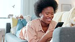 Happy young African American woman lying on sofa. Lady on furniture laughing and typing on phone. Female wearing ear piece and communicating through mobile device.