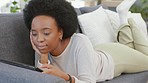 African woman scrolling on a digital tablet while lying on the couch at home. Carefree female with afro hair browsing social media, doing online shopping and enjoying her online movie subscription 