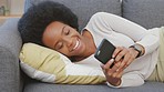Relaxed woman texting on her phone and laughing while lying on the sofa. Happy african american female browsing social media on an online app and smiling while relaxing on the couch at home