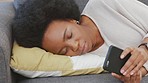 Tired black woman texting on a phone while resting on a sofa at home. Exhausted African woman falling asleep while browsing social media on a couch, using her free time to take a nap and be lazy 