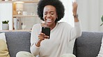 Happy African American woman expressing joy with a winning gesture while holding a phone and sitting on a sofa. Black female surprised by good news or results. Lady getting an approved business loan