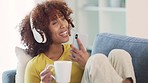 Happy woman listening to music and singing while drinking coffee at home. Cheerful young female jamming to her favorite songs and enjoying a carefree weekend while relaxing on a couch in a good mood