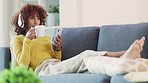 Woman taking selfies on phone, listening to music and drinking cup of coffee while relaxing on home living room sofa. Cool afro woman dancing, choosing songs from playlist and posting on social media