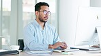 Tired and stressed man with a headache sitting alone at his desk inside an office. Exhausted businessman in pain while typing on a computer and wearing glasses, working late and looking overworked