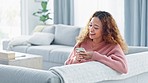 Happy woman laughing while texting on a phone and sitting on a sofa inside her living room with copy space. A cute young female enjoying online social media, looking comfortable and cheerful at home