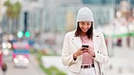 Woman laughing while reading a funny text on a phone in the city on a cold day. Portrait of a trendy and cheerful female typing a message, scrolling social media and browsing the web in a busy town