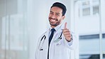 Doctor giving thumbs up hand gesture while smiling and looking confident, standing inside a hospital. Male medical professional alone showing a sign of approval and symbol of agreement and trust