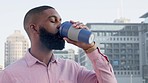 Bearded African American male drinking coffee from bamboo mug with a background of city buildings. Young employee in a smart shirt enjoying early morning hot drink. Thinking about his day before work