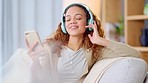 Woman wearing headphones and using a phone to listen to her music while relaxing on a sofa at home. Carefree female dancing and having fun alone on a couch, enjoying her free time on a weekend