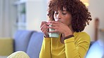 Trendy woman enjoying coffee and blowing to cool it down while relaxing at home. African female with curly afro holding and drinking hot cup while thinking and daydreaming in her cozy apartment