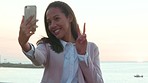 Trendy tourist taking a selfie on her phone by the sea with clear sky copy space. Young vlogger exchange student by the ocean, taking a portrait photo with emoji hand gesture for her social media app